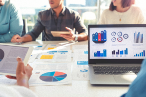 Group of people meeting with technology and paperwork. Close up of digital tablet and laptop computer with market analysis data on the screen. The documents have financial or marketing figures, graphs and charts on them. Multi ethnic group including Caucasian, latin and African American